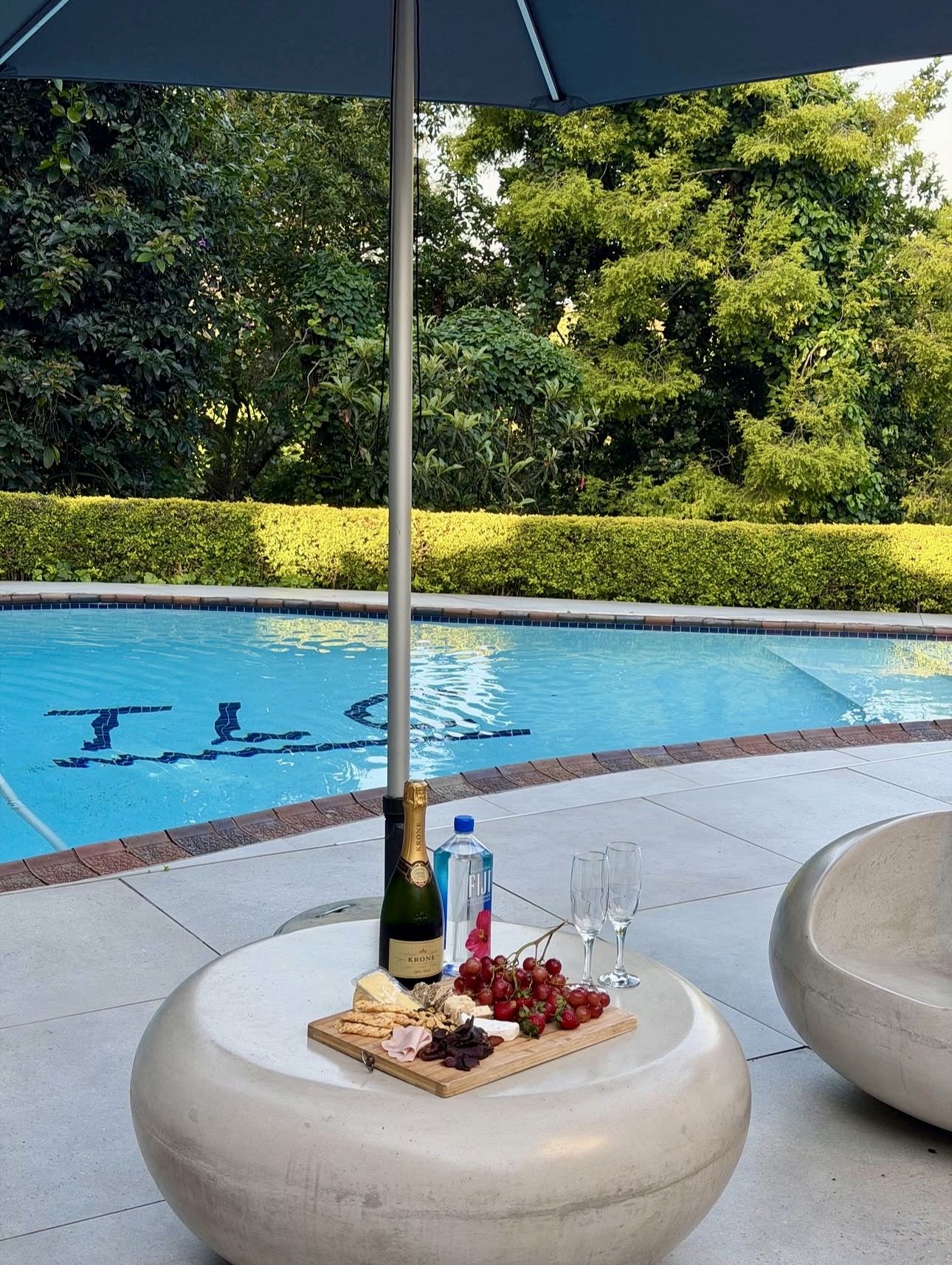 A cheese board and a bottle of bubbly on a stone table by the pool