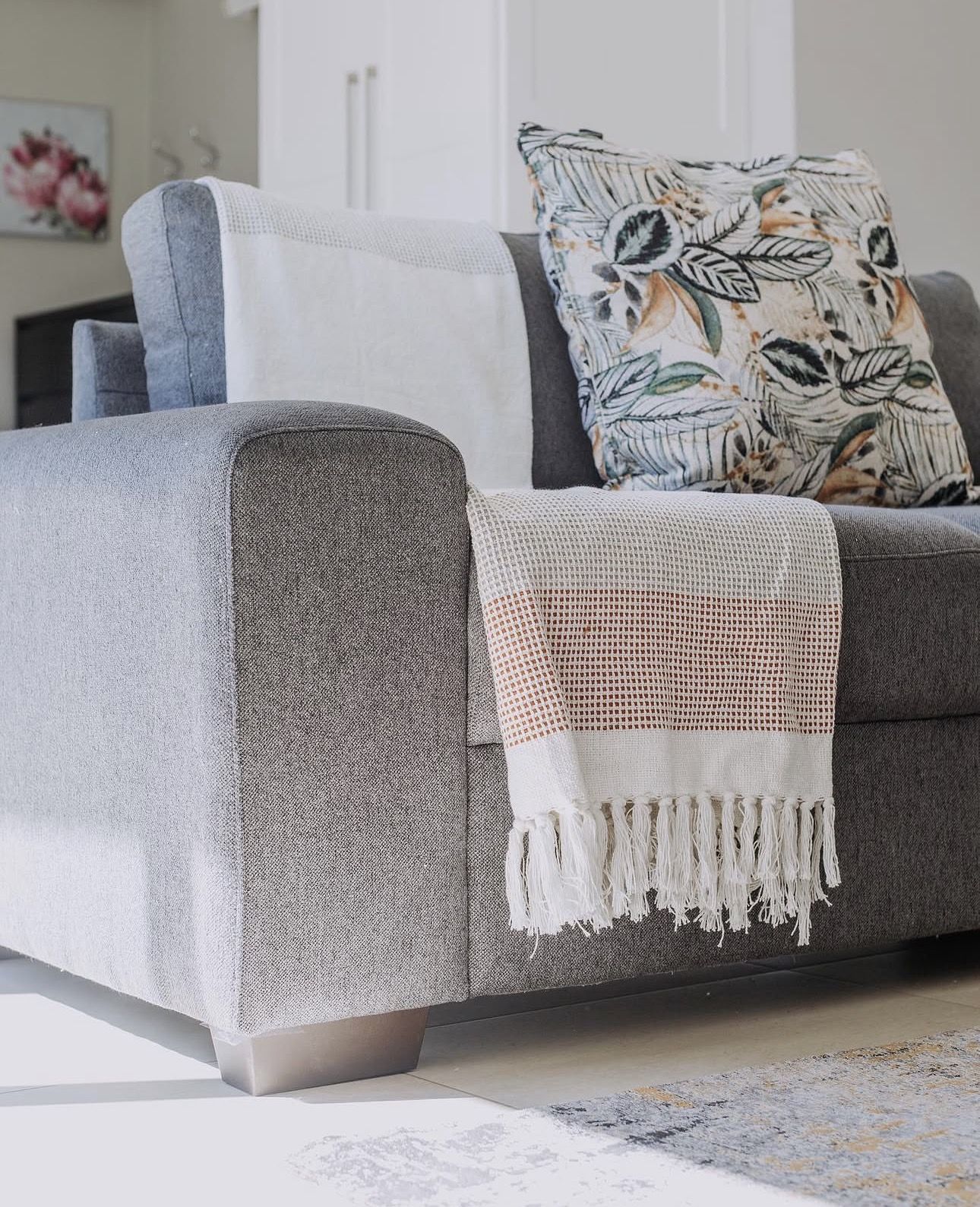 A grey couch with a leaf print cushion and a fringed throw