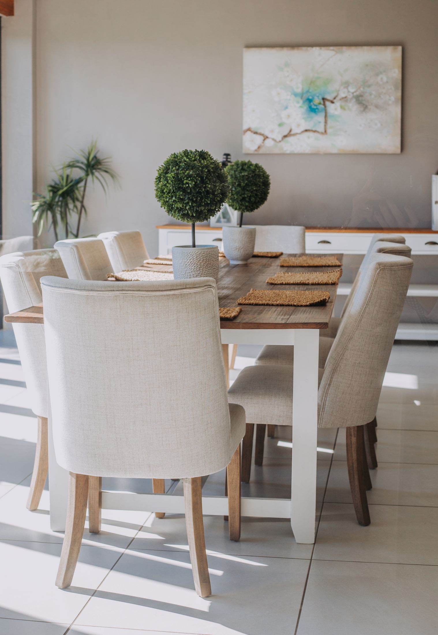 The Unit 5 dining table with linen chairs and two potted topiaries