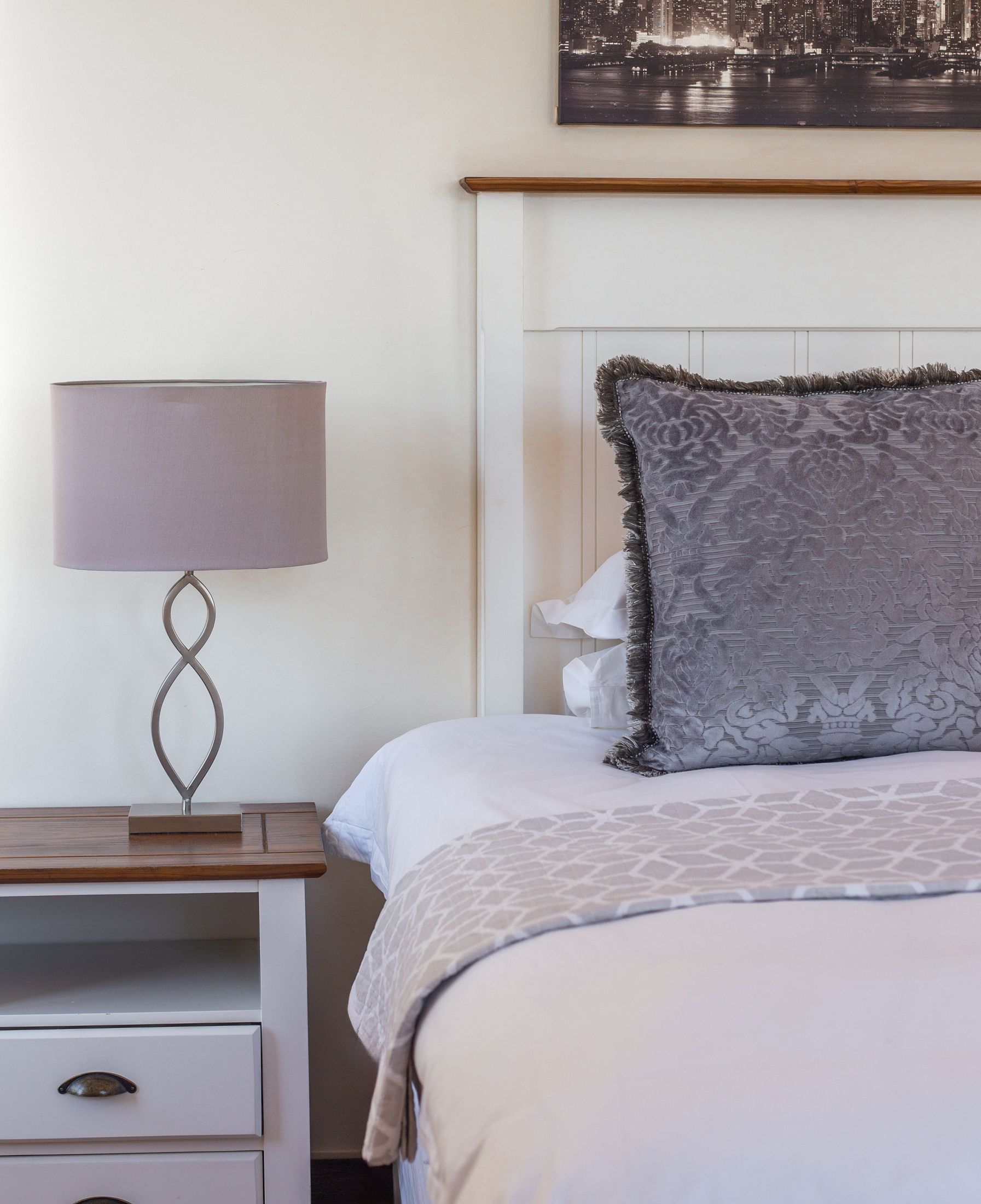 A grey cushion and a lamp beside a bed with a white headboard