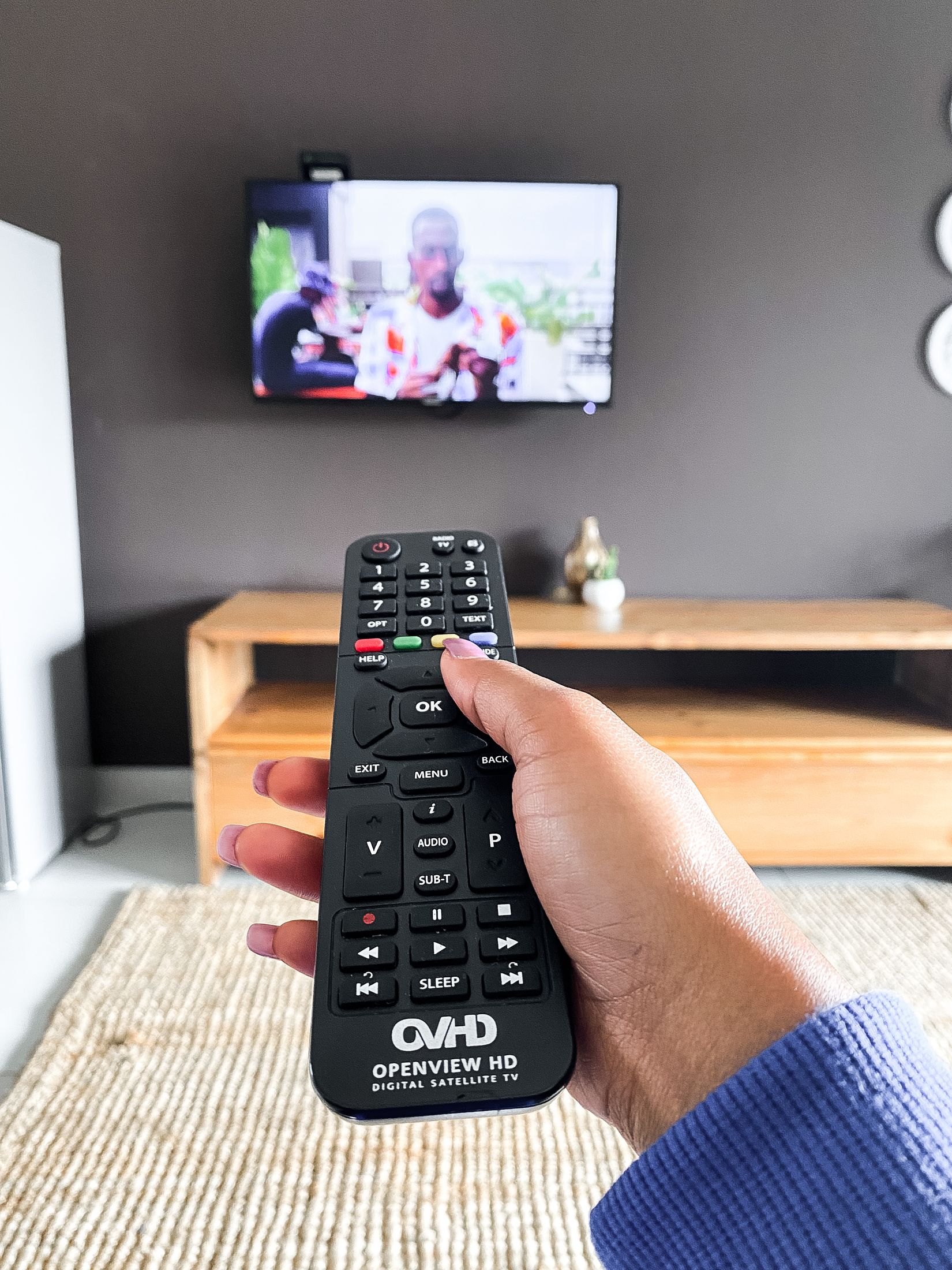 A hand holding the TV remote in front of the lounge TV
