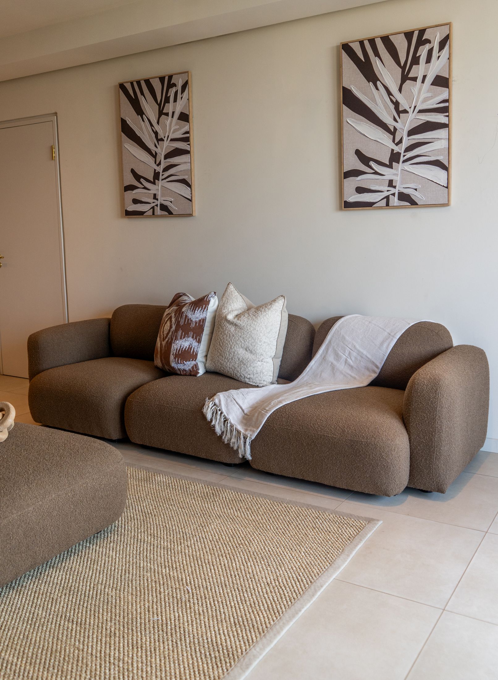 A brown curved couch with cushions and a white throw under two leaf prints